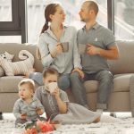 Happy family with children spending time at home while parents sitting on couch with cups of coffee and looking at each other when kids resting on floor
